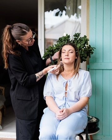 Laura maquille une cliente assise devant des volets turquoise